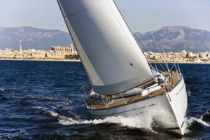 Yacht a vela bianco naviga sull'acqua con vele spiegate e paesaggio montano sullo sfondo.
