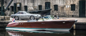 Barca Riva in legno con dettagli bianchi ormeggiata accanto a un'auto sportiva grigia in un porto.